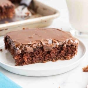 A slice of chocolate sheet cake with chocolate icing on a white plate, with a baking tray and a glass of milk in the background.