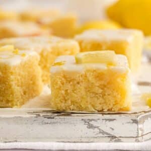 Close-up of lemon brownies topped with glaze and small lemon pieces, arranged on a rustic white surface.