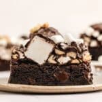 A close-up of a chocolate brownie topped with marshmallows, chopped nuts, and chocolate drizzle on a ceramic plate.