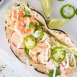 A shrimp taco on a corn tortilla topped with sliced radishes, jalapeños, shredded cabbage, cilantro, and a drizzle of white sauce, with lime wedges nearby.