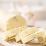 Sliced log of herb butter on parchment paper, showing visible pieces of green herbs inside the creamy butter.