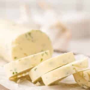 Sliced log of herb butter on parchment paper, showing visible pieces of green herbs inside the creamy butter.