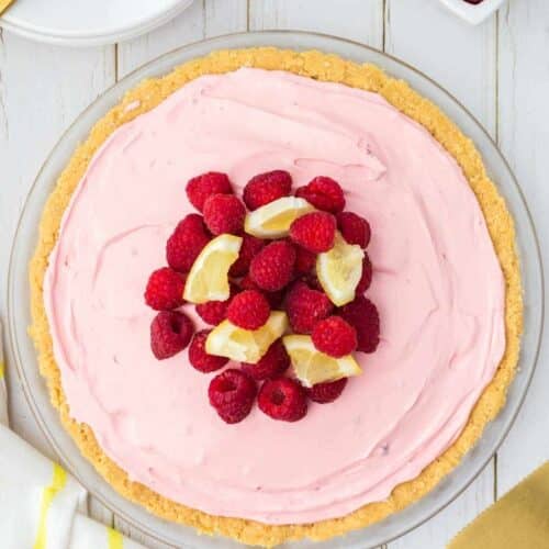 A pie with a pink creamy filling in a crumb crust, topped with fresh raspberries and small lemon wedges, on a white wooden surface.