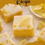 Several lemon brownies with white icing and lemon slices are arranged on parchment paper, with a plate of additional lemon slices in the background.