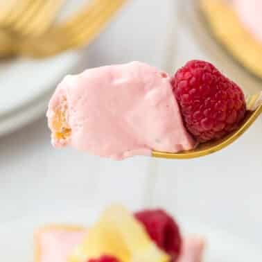 A fork holds a bite of pink creamy dessert with a fresh raspberry on top, against a blurred background of plates and forks.