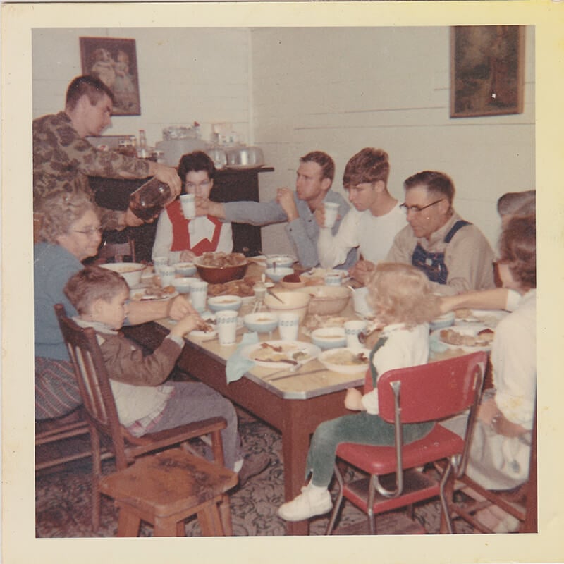 Nine people of various ages sit around a dining table sharing a meal, with one person pouring coffee. The table is set with dishes and food in a homey setting.