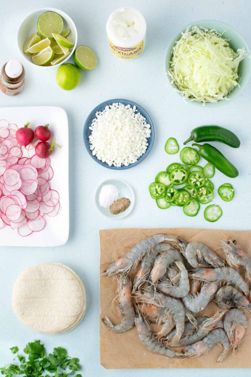 Ingredients for shrimp tacos arranged on a surface, including raw shrimp, tortillas, radish slices, jalapeños, cabbage, cotija cheese, lime, spices, crema, and cilantro.