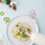 A taco on a white plate is topped with radish slices, cilantro, and sauce being poured from a squeeze bottle; Tajin seasoning and jalapeño slices are nearby.