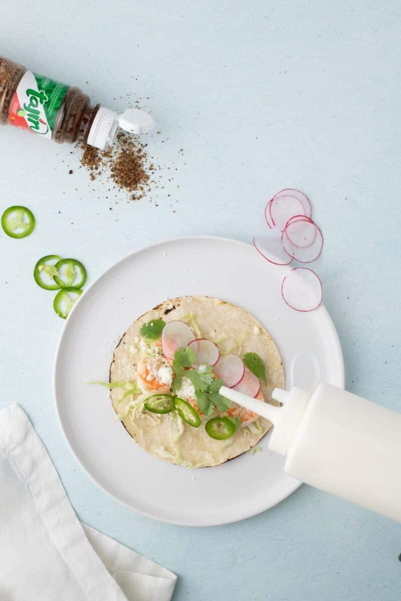 A taco on a white plate is topped with radish slices, cilantro, and sauce being poured from a squeeze bottle; Tajin seasoning and jalapeño slices are nearby.