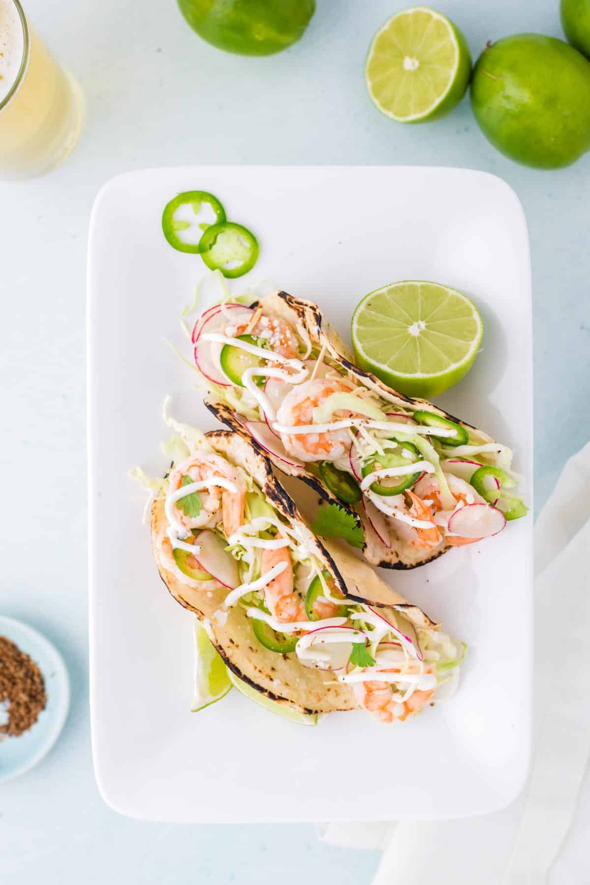 Two baja shrimp tacos with shredded cabbage, radish, jalapeño slices, and sauce on corn tortillas are served on a white plate with a halved lime.