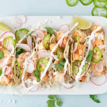 A rectangular white plate with four baja  shrimp tacos topped with sliced radishes, jalapenos, cilantro, cheese, and drizzled sauce, garnished with lime wedges.