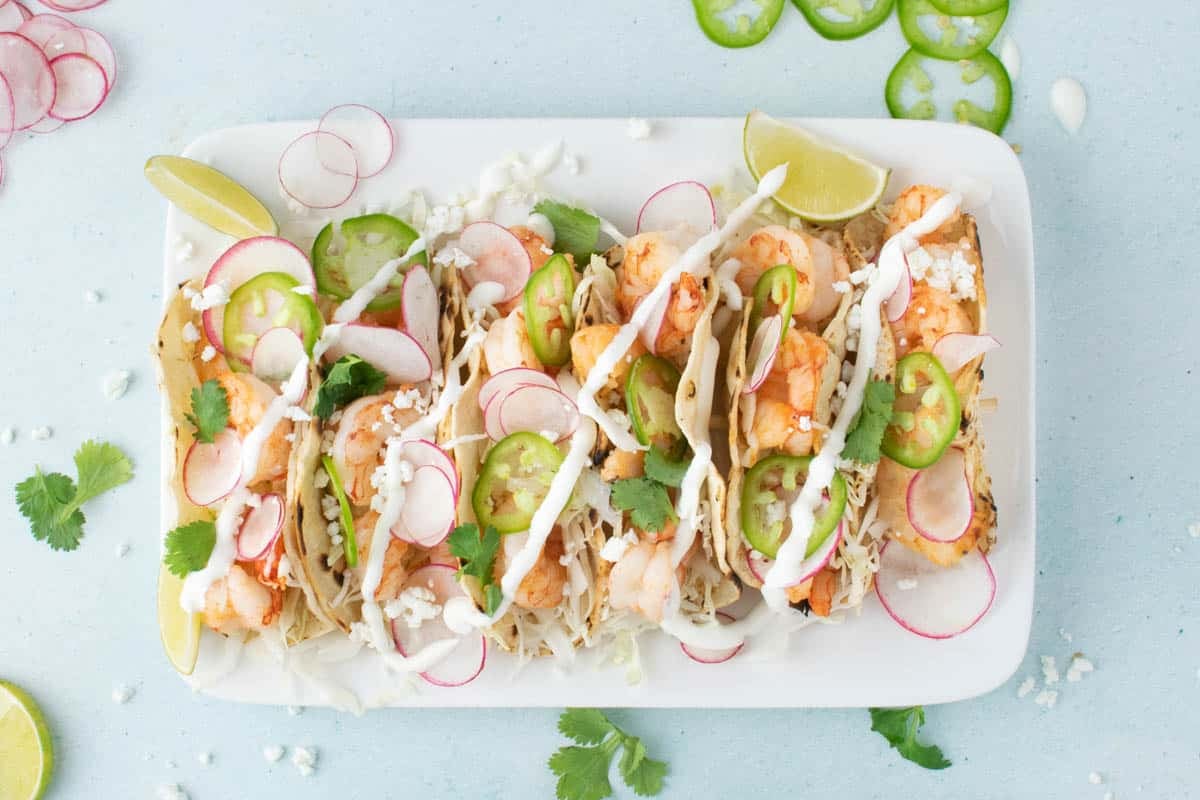 A rectangular white plate with four baja  shrimp tacos topped with sliced radishes, jalapenos, cilantro, cheese, and drizzled sauce, garnished with lime wedges.