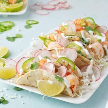 A rectangular white plate with shrimp tacos topped with sliced radishes, jalapenos, cilantro, and crema, garnished with lime wedges on a light blue surface.