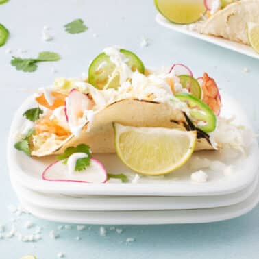 A taco filled with shrimp, shredded cabbage, sliced radish, jalapeno, and cilantro is served on a plate with a lime wedge and a drizzle of white sauce.