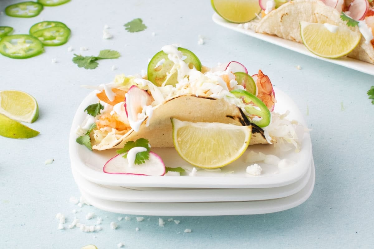 A taco filled with shrimp, shredded cabbage, sliced radish, jalapeno, and cilantro is served on a plate with a lime wedge and a drizzle of white sauce.