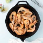 A cast iron skillet filled with cooked shrimp, next to a white napkin, metal tongs, half a lime, and a small bottle of oil on a light surface.