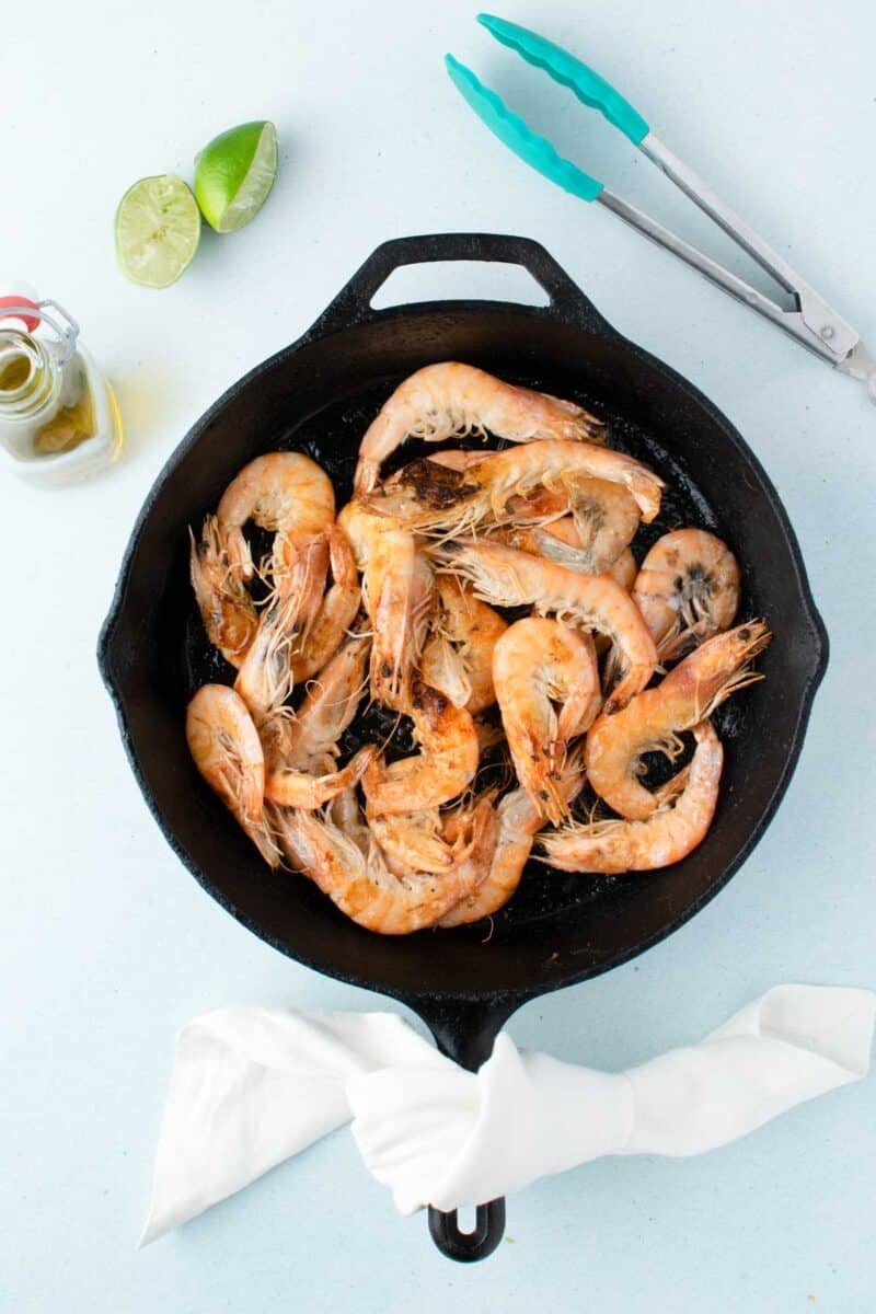 A cast iron skillet filled with cooked shrimp, next to a white napkin, metal tongs, half a lime, and a small bottle of oil on a light surface.