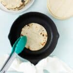 A person uses tongs to heat a corn tortilla on a cast iron skillet, with a stack of tortillas and a bowl of cooked tortillas nearby.