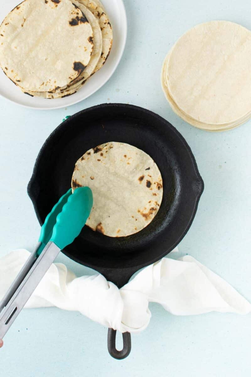 A person uses tongs to heat a corn tortilla on a cast iron skillet, with a stack of tortillas and a bowl of cooked tortillas nearby.