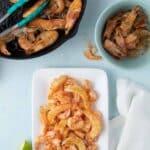Cooked shrimp in a skillet with tongs, shrimp shells in a bowl, and peeled shrimp arranged on a white plate with a lime wedge nearby on a light blue surface.