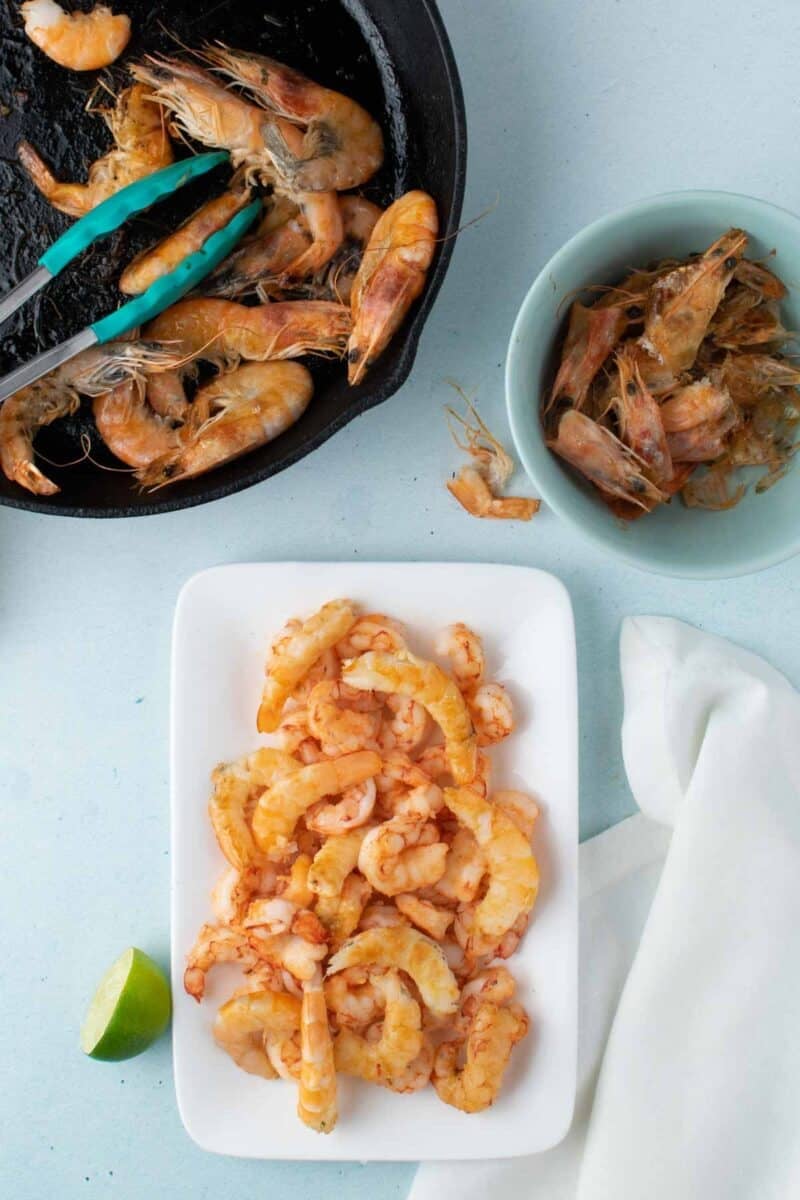 Cooked shrimp in a skillet with tongs, shrimp shells in a bowl, and peeled shrimp arranged on a white plate with a lime wedge nearby on a light blue surface.