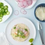 A tortilla on a plate topped with shrimp, radish slices, jalapeños, and cilantro. Surrounding dishes hold radish slices, jalapeño slices, cheese, and fresh cilantro.
