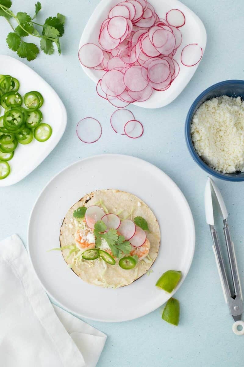 A tortilla on a plate topped with shrimp, radish slices, jalapeños, and cilantro. Surrounding dishes hold radish slices, jalapeño slices, cheese, and fresh cilantro.