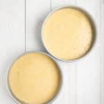 Two round metal cake pans filled with yellow cake batter are placed on a white wooden surface.