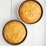 Two round golden-brown cakes in metal pans are placed on a white wooden surface.