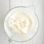 A glass mixing bowl with whipped cream or frosting on a white wooden surface.