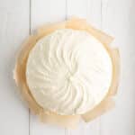 A round cake with smooth white frosting sits on parchment paper, placed on a white wooden surface.