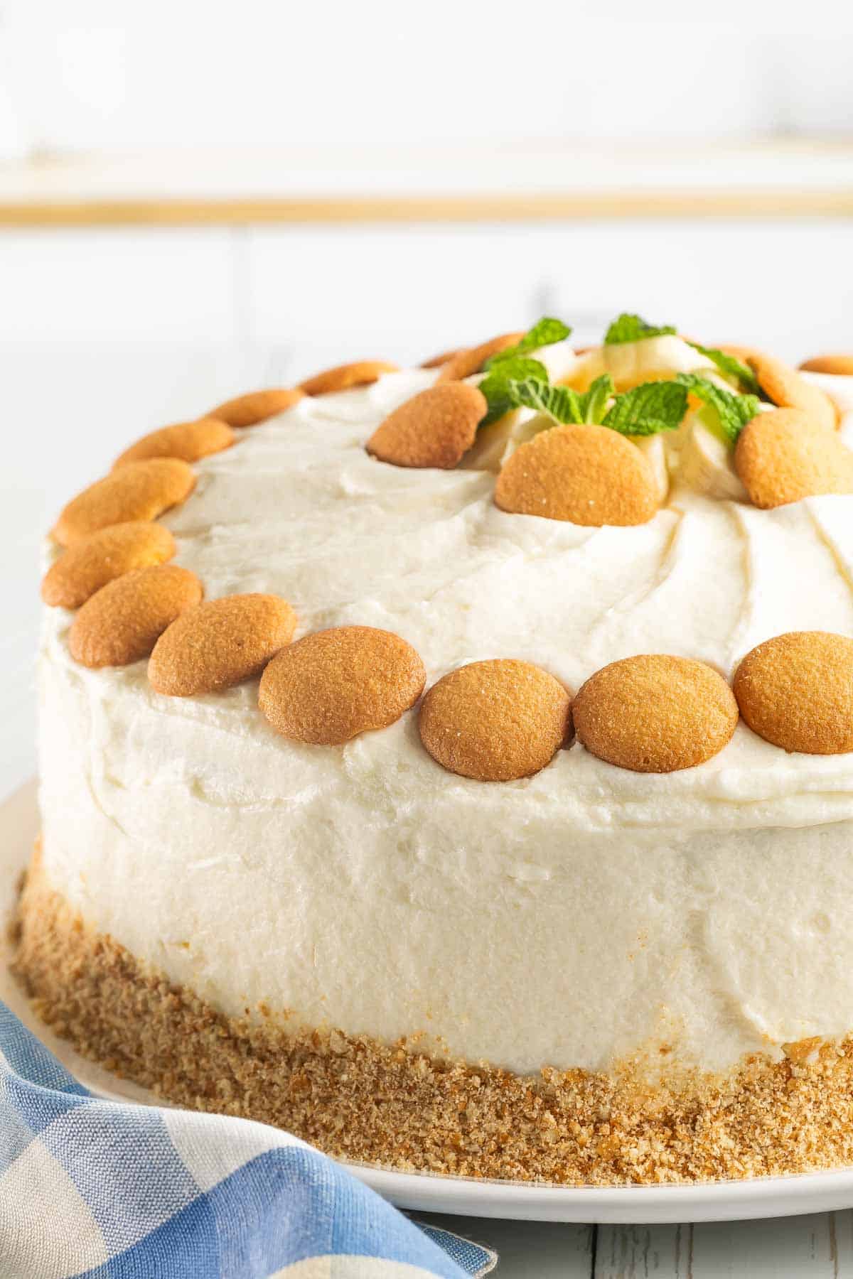 A banana pudding layer cake with white frosting, decorated with vanilla wafer cookies on top and around the base, garnished with mint leaves.