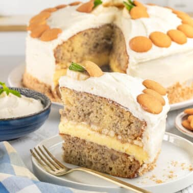 A slice of layered banana cake with frosting and vanilla wafers is served on a plate, with the rest of the cake, a bowl of frosting, and extra wafers in the background.