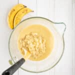 A glass bowl with mashed bananas and batter being mixed with a spatula sits on a white wooden surface next to two ripe bananas.
