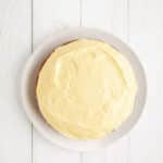 A round cake with yellow frosting is placed on a white oval plate, set on a white wooden surface.