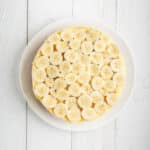 A round cake topped with evenly sliced bananas, placed on a white plate over a white wooden surface.