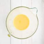 A glass mixing bowl containing a yellow liquid mixture sits on a white wooden surface.