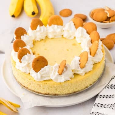 Banana cheesecake topped with whipped cream and vanilla wafer cookies, with bananas and extra cookies in the background on a light surface.