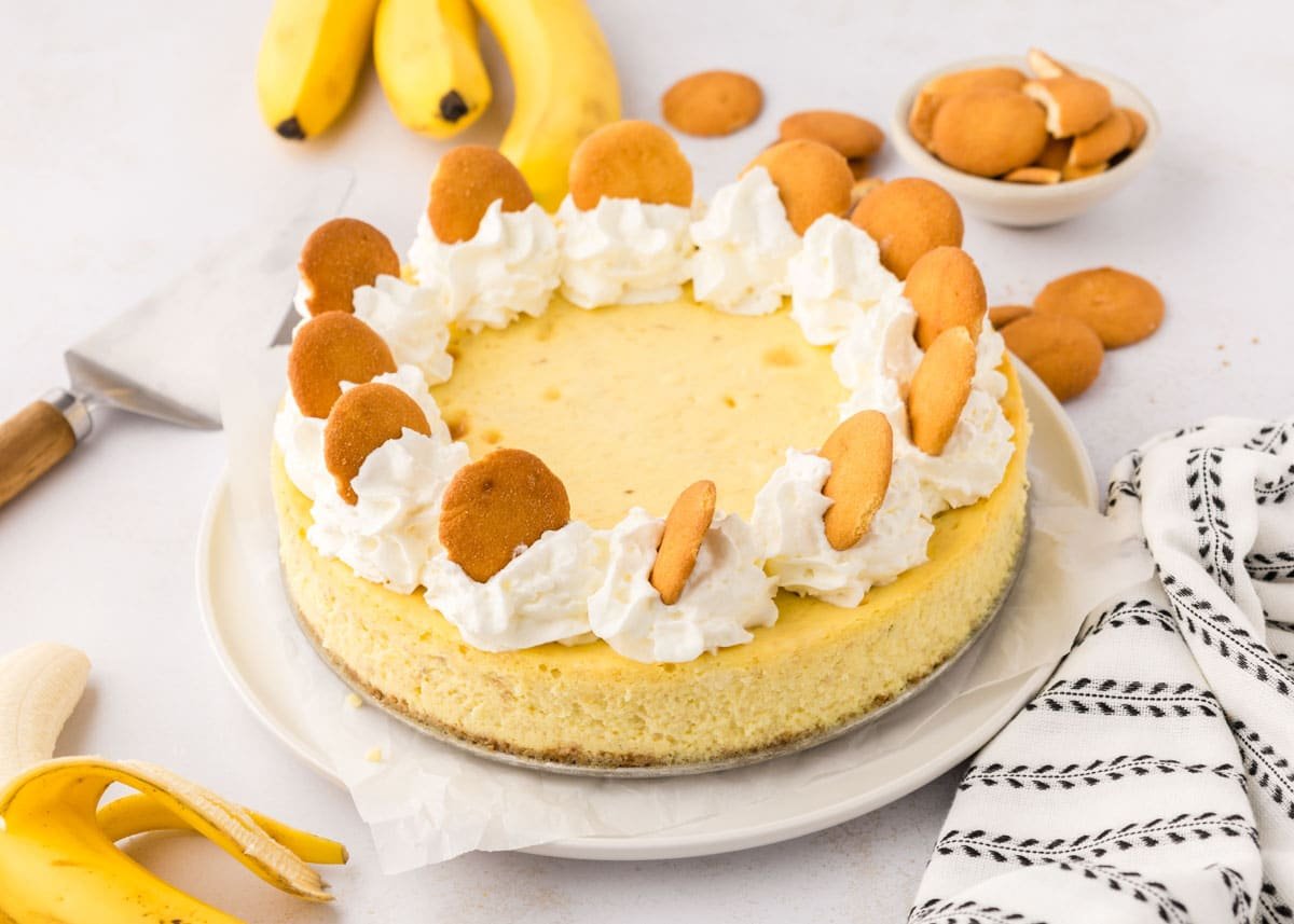 Banana cheesecake topped with whipped cream and vanilla wafer cookies, with bananas and extra cookies in the background on a light surface.