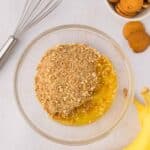 A glass bowl with crushed cookies and melted butter on a white surface, next to a whisk, whole cookies, and two bananas.
