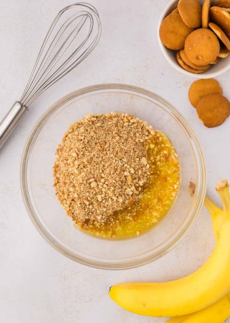A glass bowl with crushed cookies and melted butter on a white surface, next to a whisk, whole cookies, and two bananas.