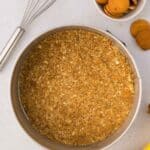 A springform pan filled with a crumbly cookie crust sits on a countertop next to a whisk, a bowl of vanilla wafers, and a banana.