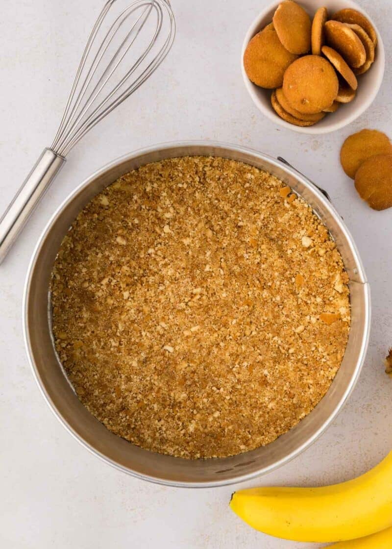 A springform pan filled with a crumbly cookie crust sits on a countertop next to a whisk, a bowl of vanilla wafers, and a banana.