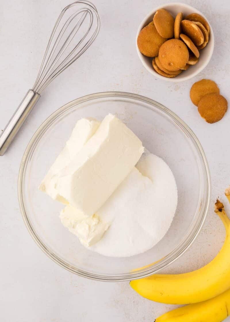 A glass bowl with cream cheese and sugar, a whisk, a bowl of vanilla wafers, and two bananas on a light surface.