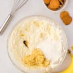 A glass bowl contains whipped cream cheese, mashed banana, sugar, and vanilla extract. Nearby are a whisk, vanilla wafer cookies, and whole bananas on a white surface.