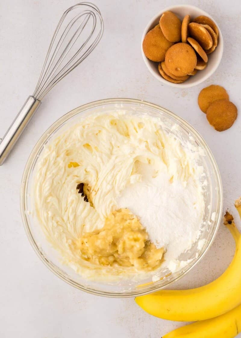 A glass bowl contains whipped cream cheese, mashed banana, sugar, and vanilla extract. Nearby are a whisk, vanilla wafer cookies, and whole bananas on a white surface.