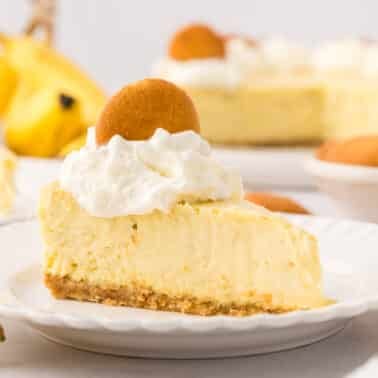 A slice of banana cream cheesecake topped with whipped cream and a vanilla wafer, served on a white plate with bananas and more cheesecake in the background.