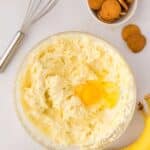 A glass bowl of creamy batter with a raw egg on top, next to a metal whisk, a bowl of vanilla wafers, and two bananas on a white surface.