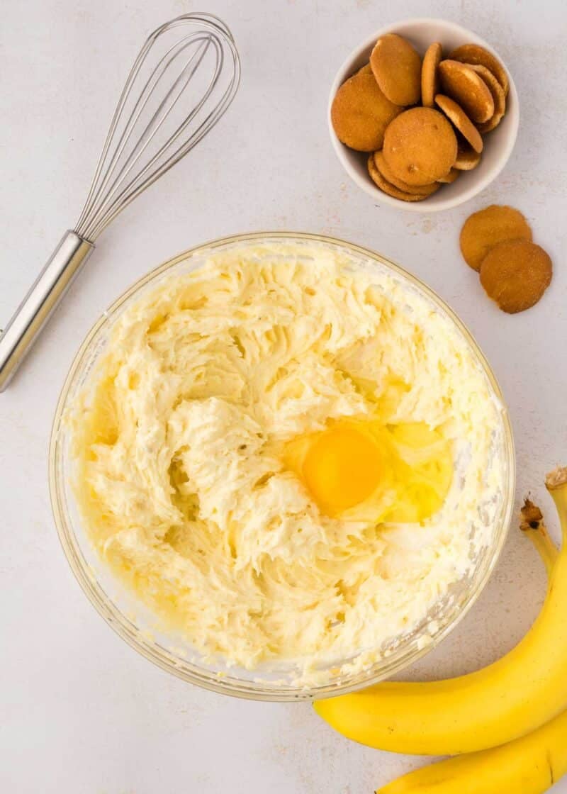 A glass bowl of creamy batter with a raw egg on top, next to a metal whisk, a bowl of vanilla wafers, and two bananas on a white surface.
