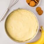 A mixing bowl of banana pudding with a whisk, a bunch of bananas, and a small bowl of vanilla wafers on a white surface.
